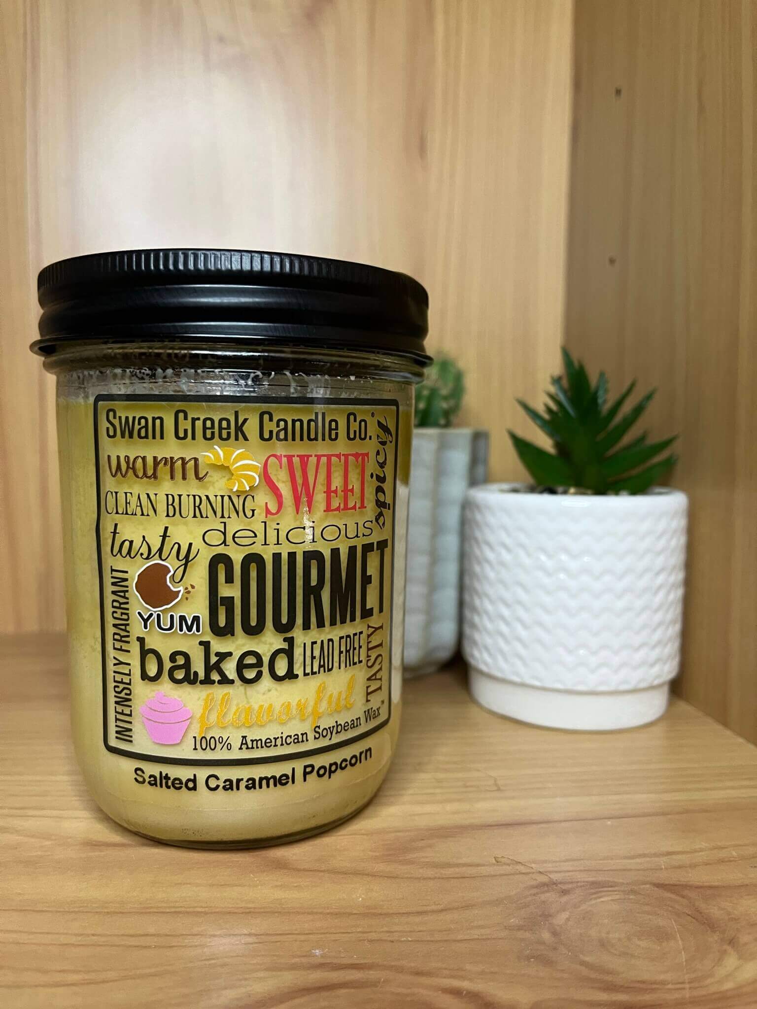 A photo of a golden brown candle on a wooden shelf with a mini plant in a white pot in the background